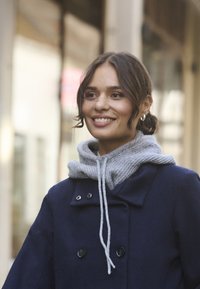 Mujer sonriente con cabello oscuro recogido en un moño bajo que lleva pendientes de aro plateados, un gorro de punto gris y un abrigo azul marino, mientras está de pie al aire libre.