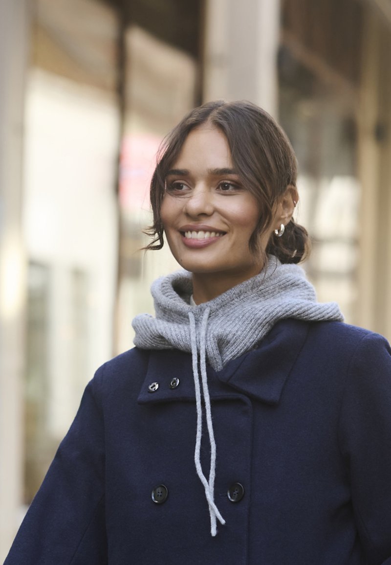 Mujer sonriente con cabello oscuro recogido en un moño bajo que lleva pendientes de aro plateados, un gorro de punto gris y un abrigo azul marino, mientras está de pie al aire libre.