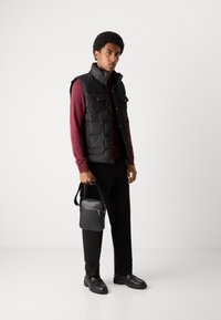 Čierna prešívaná vesta s prednými vreckami navlečená na vínovom svetri, v kombinácii s čiernymi nohavicami a robustnými čiernymi topánkami, s čiernym crossbody batohom.