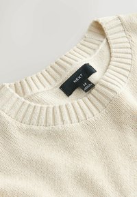 Roomkleurige gebreide trui met geribde halslijn en zwart maatlabel met de tekst "NEXT M Made in Bangladesh."