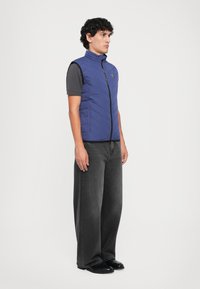 Jovem em pé, vestindo calças largas pretas, sapatos pretos, t-shirt cinza escura e um colete azul acolchoado sem mangas.