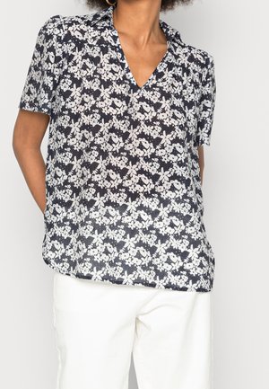 Blouse à manches courtes en marine avec un motif floral blanc, décolleté en V, tissu texturé et coupe décontractée. Associée à un pantalon de couleur claire.