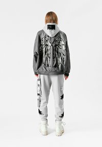 Bomber gris con bordado de alas plateadas en la parte trasera, combinado con pantalones de chándal grises con detalles en negro y gráficos, usados con botas blancas.