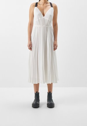 Femme portant une robe midi plissée blanche avec des bretelles noires et des bottines noires épaisses, debout sur un fond blanc uni.