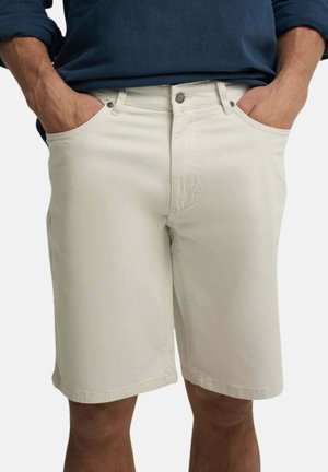 Hombre con pantalones cortos beige hasta la rodilla y una camisa azul oscuro de manga larga con las manos en los bolsillos sobre un fondo blanco.