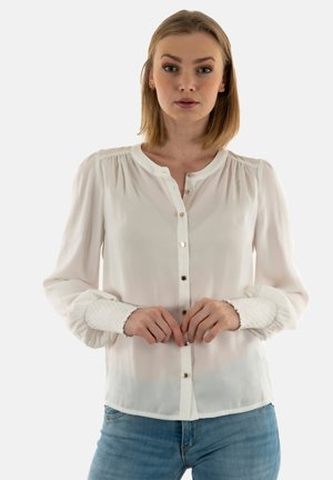 Blouse blanche à boutons avec des épaules froncées et des poignets texturés contrastants. Elle présente un tissu doux et transparent, avec six boutons sur le devant.