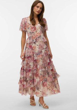 Kvinde i en lagdelt, blomstret maxikjole med pink, lilla og beige nuancer, stående mod en ensfarvet hvid baggrund.