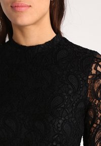 Haut en dentelle noire avec un col montant, des motifs cachemire délicats et des manches en dentelle transparente, mettant en valeur une apparence texturée.