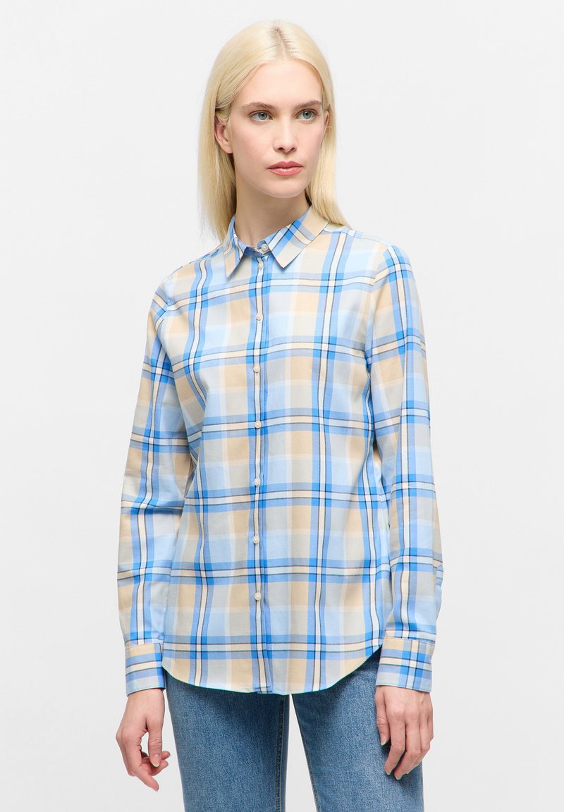 Frau trägt ein karierter Hemd in Blau, Beige und Weiß, sowie eine blaue Jeans, und steht vor einem einfarbigen hellen Hintergrund.