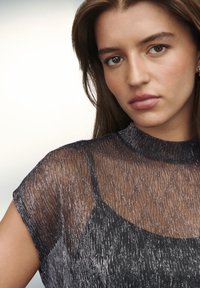 Läbipaistev must pluus kõrge kaelusega, millel on metallilised niidid ja tekstuuriga muster. Lühikesed varrukad ja keha järgnev siluett.