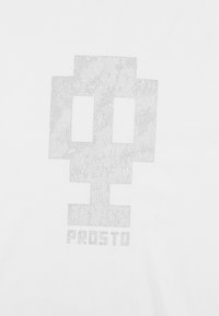 Biała koszulka z dużym, odblaskowym, srebrnym wzorem w kształcie czaszki z napisem "PROSTO" poniżej. Gładka, miękka tekstura.