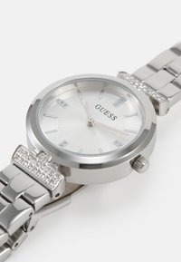 Montre en acier inoxydable argenté avec un cadran rond, un cadran texturé blanc, des marqueurs d'heure en cristal et un bracelet à maillons. Agrémentée de détails en strass.
