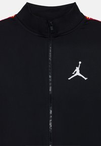 Veste zippée noire en tissu lisse, dotée d'un col haut, de accents d'épaule rouges et jaunes, et d'un logo Jumpman blanc sur la poitrine.