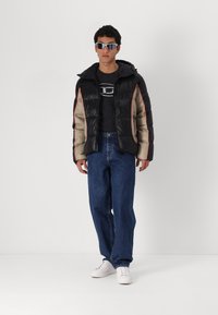 Casaco acolchoado preto e castanho claro com detalhes em vermelho, combinado com uma t-shirt preta, jeans azuis e ténis brancos.