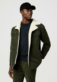 Grüne Cordjacke mit einem cremefarbenen Kunstfellkragen, Fronttaschen, kombiniert mit einem dunkelblauen Pullover und passenden grünen Hosen. Schwarze Mütze.