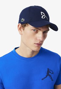 Kepala baseball navy dengan pinggiran melengkung dan logo putih, dipadukan dengan kaos lengan pendek biru yang menampilkan cetakan grafis gelap.