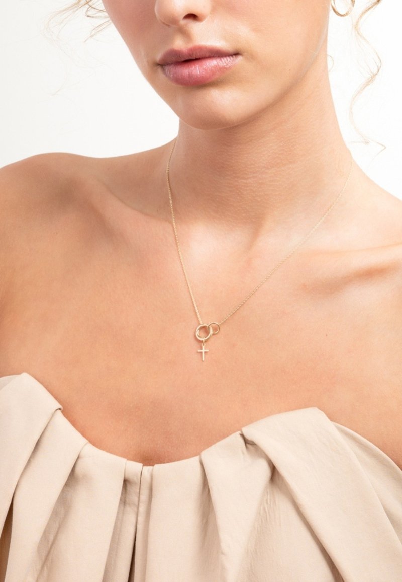 Gouden halsketting met in elkaar grijpende ringen en een klein kruisjesspeldje. De kettingdraadjes zijn delicaat, wat een gladde huidoppervlakte benadrukt. Beige stof hangt eronder.