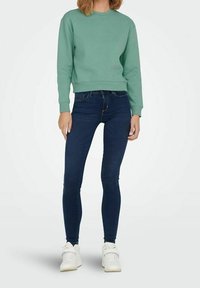 Osoba v mátově zelené mikině, tmavě modrých skinny džínech a bílých teniskách, stojící proti jednoduchému světlému pozadí.