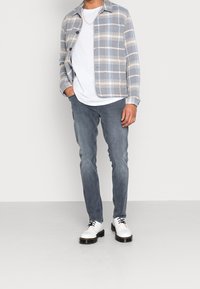 Camisa de cuadros en azul y beige, camiseta blanca, vaqueros grises y zapatos blancos con suelas negras. Presenta cierre de botones en la parte delantera y cuello.