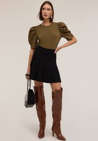 Top oliva de mangas abullonadas, falda corta negra con pliegues, botas marrones hasta la rodilla y un bolso negro con cadena.