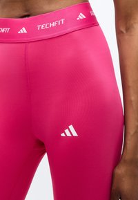 Κοντινό πλάνο ενός ατόμου που φορά φωτεινά ροζ ελαστικά κολάν Adidas Techfit με λογότυπα στη μέση και στο μηρό.