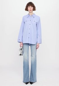 Camicia azzurro chiaro con bottoni e due tasche sul petto, abbinata a jeans a campana in denim chiaro e una borsa argentata, indossata da un modello su uno sfondo bianco.