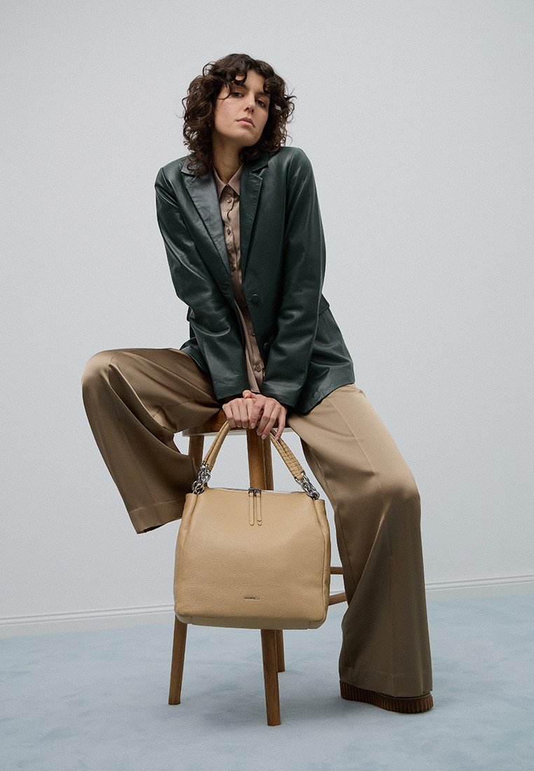 Sieviete ar viļņotiem matiem, ģērbusies tumši zaļā žaketē un bēšu platā kāju biksēs, sēž uz koka krēsla, tur bēšu roku somu.