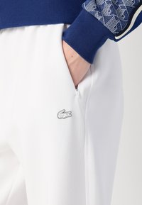 Vita sweatpants med en dragkedjeficka, med en liten broderad krokodil-logotyp. Toppen har blå ribbade manchetter och geometriska mönster.