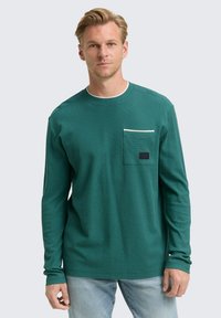 Blonde man in een groene langemouw shirt met een borstzak en lichtblauwe jeans, staand tegen een effen lichte achtergrond.