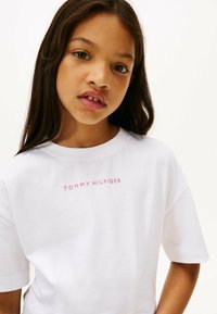 Biała bawełniana koszulka z okrągłym dekoltem i krótkimi rękawami, z nadrukiem "TOMMY HILFIGER" w jasnoróżowym kolorze na klatce piersiowej.