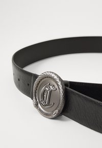 Svart lærbelte med en rund sølvmetallspenne med slange motiv og inngravert "Just Cavalli" tekst; glatt tekstur og klassisk design.