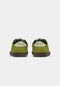 Sapatilhas verdes de camurça com uma sola de borracha castanha texturizada. Gola branca acolchoada com a marca "VEJA" na parte traseira. Design minimalista.
