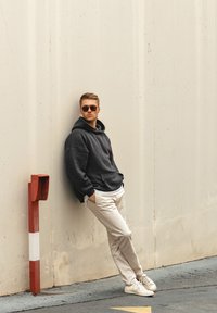 Felpa nera oversize, pantaloni beige chiaro e sneaker bianche. In piedi contro un muro, indossa occhiali da sole. Design semplice con una vestibilità casual.