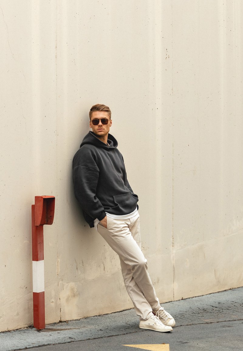 Felpa nera oversize, pantaloni beige chiaro e sneaker bianche. In piedi contro un muro, indossa occhiali da sole. Design semplice con una vestibilità casual.