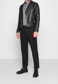 Giacca di pelle nera da biker con cerniere argento, camicia grigia a bottoni, pantaloni neri e stivali neri alla caviglia con suola texturizzata.