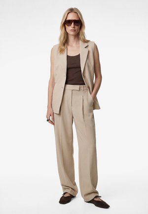 Frau mit beige ärmellosem Blazer und weit geschnittenen Hosen, braunem Tanktop, dunkelbraunen Ballerinas und übergroßen Schildpatt-Sonnenbrillen, stehend.