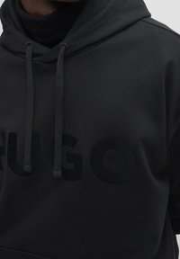 Sudadera negra con una tela texturizada, que presenta un gran logo tonal de "HUGO". Incluye capucha con cordón ajustable y puños acanalados.