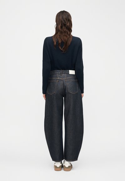 Γυναίκα με μακριά κυματιστά μαλλιά, ντυμένη με μαύρο μακρυμάνικο τοπ, σκούρο τζιν παντελόνι με φαρδύ πόδι και λευκά αθλητικά παπούτσια, γυρισμένη πλάτη σε απλό φόντο.