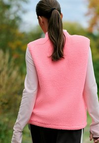 Svetlo roza brezrokavnik iz flisa z teksturirano površino, brez rokavov, visok ovratnik, nošen čez dolgo majico z dolgimi rokavi, pogled od zadaj.
