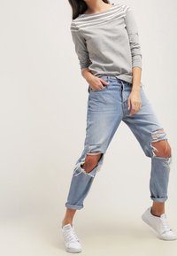 Un haut à manches longues rayé gris et blanc associé à un jean bleu clair distendu, avec des genoux déchirés et des ourlets retroussés, le tout complété par des baskets blanches.