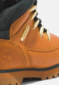 Bota de cuero marrón con un cuello negro, hardware dorado y el prominente logo de Timberland. Presenta una suela resistente con patrones de pisada claramente definidos.