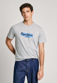 Siva pamučna majica s plavim i crnim logotipom "Pepe Jeans London", u kombinaciji s tamnim trapericama. S kratkim rukavima i okruglim izrezom.