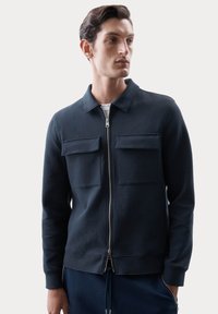 Giacca zip-up navy con due tasche sul petto, polsini a costine e colletto piatto, realizzata in un tessuto morbido e strutturato.