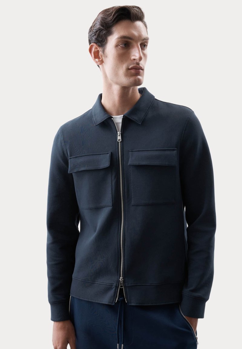 Giacca zip-up navy con due tasche sul petto, polsini a costine e colletto piatto, realizzata in un tessuto morbido e strutturato.