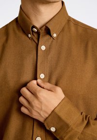 Bruine button-up overhemd met een kraag, voorzien van lichtgekleurde knopen en een gestructureerde stof, gedeeltelijk dichtgeknoopt aan de voorkant.
