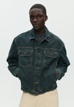 Persona con cabello corto que lleva una chaqueta de mezclilla oscura con bolsillos abotonados y pantalones de color claro, posando frente a un fondo liso.