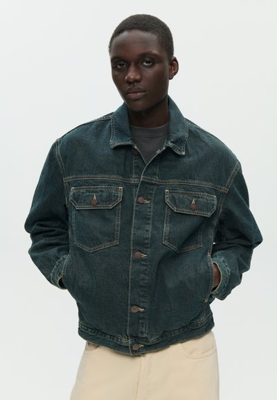 Persona con capelli corti che indossa una giacca di jeans scura con tasche con bottoni e pantaloni di colore chiaro, in piedi davanti a uno sfondo semplice.