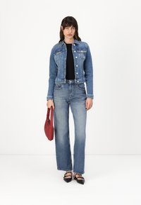 Giacca di jeans blu con bottoni, abbinata a jeans a vita alta leggermente svasati. Top nero e borsa rossa. Scarpe nere a punta.