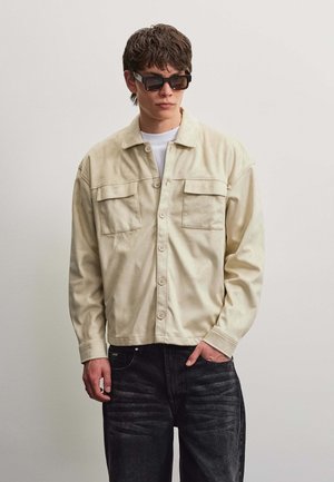 Jeune homme portant une veste crème boutonnée avec deux poches poitrine, lunettes de soleil noires, chemise blanche et jean foncé, debout devant un fond uni.