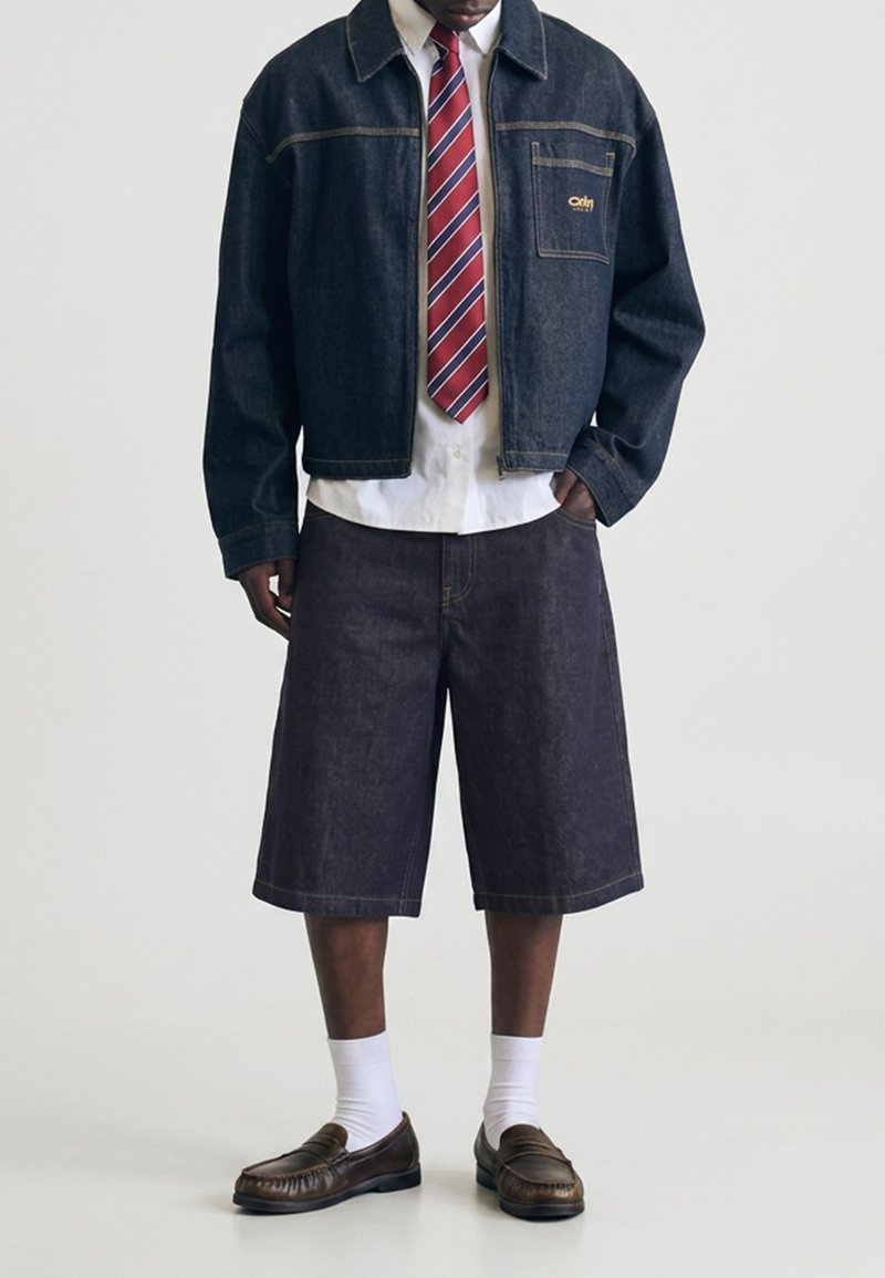 Homme portant une veste en jean foncé et un short long assorti avec une chemise blanche, une cravate rouge à rayures, des chaussettes blanches et des mocassins marron.